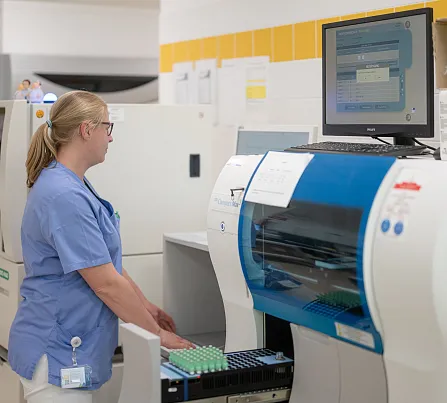 Eine Frau in blauer Arbeitskleidung steht in einem Labor vor einem medizinischen Analysegerät und bedient einen Computer. Regale sind im Hintergrund zu sehen.