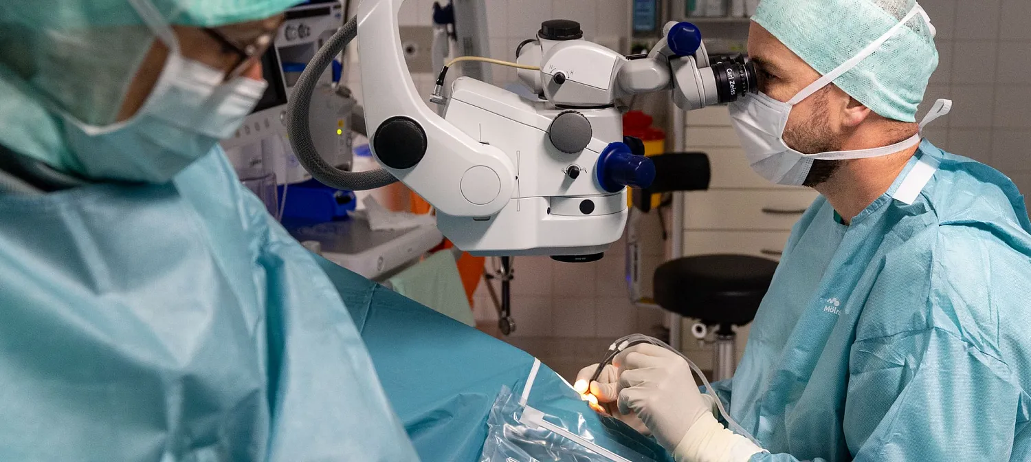 Zwei Chirurgen in OP-Kleidung führen eine mikrochirurgische Operation mit einem Mikroskop durch. Sie arbeiten sorgfältig in einem sterilen Operationssaal.