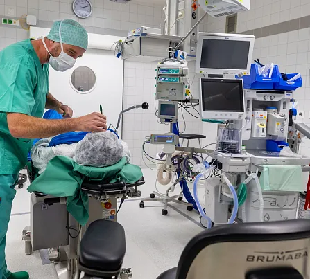 Ein Operationssaal mit einem Chirurgen in grüner OP-Kleidung, der eine Person auf einem OP-Tisch vorbereitet. Umgeben von medizinischen Geräten und Monitoren.