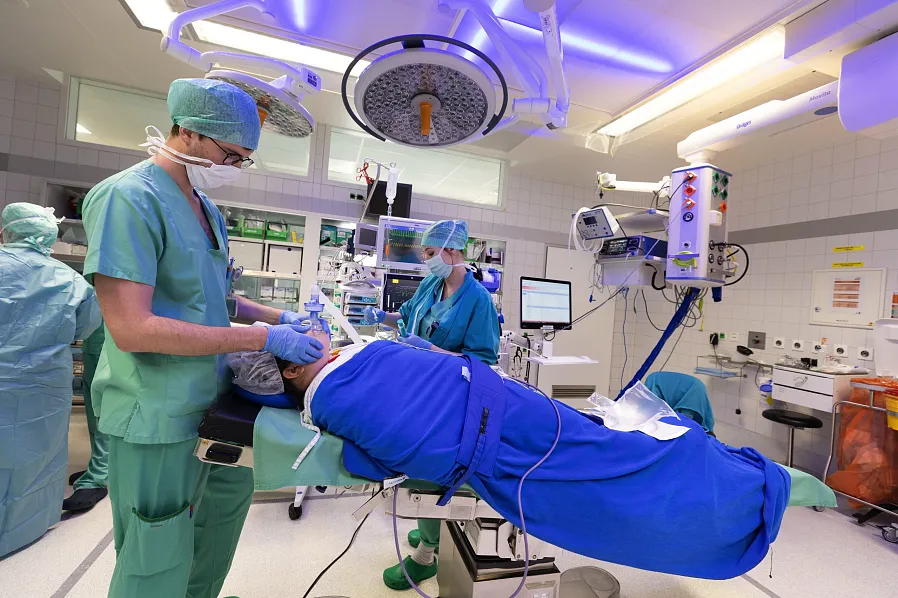 Medizinisches Team in einem Operationssaal bereitet sich auf eine Operation vor. Chirurgen tragen grüne OP-Kleidung und Masken, ein Patient liegt auf dem Tisch.