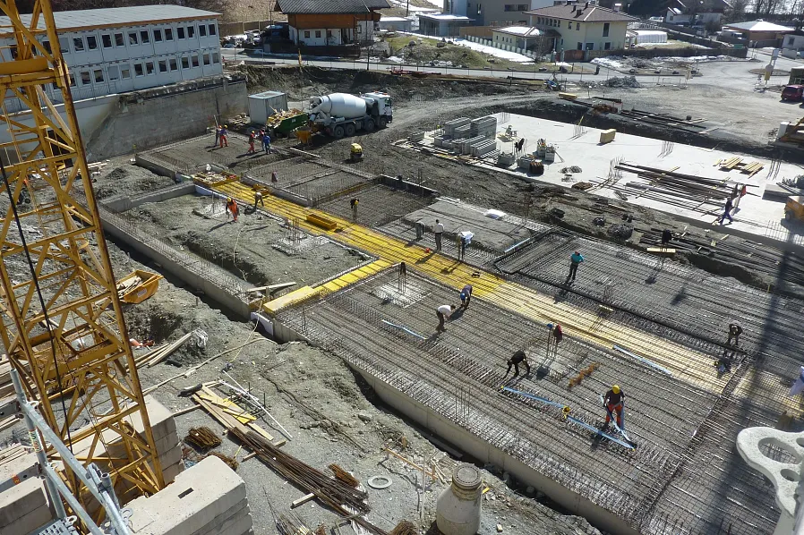 Eine Baustelle mit mehreren Arbeitern, die Stahlträger auf einem breiten, betonierten Bereich verlegen. Ein gelber Baukran ist links zu sehen, Gebäude im Hintergrund.