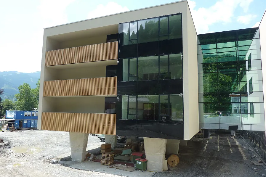 Modernes Gebäude mit großen Glasfenstern und Holzbalkonen auf Stelzen. Im Hintergrund sind Berge und Wolken zu sehen, der Boden ist unbefestigt.
