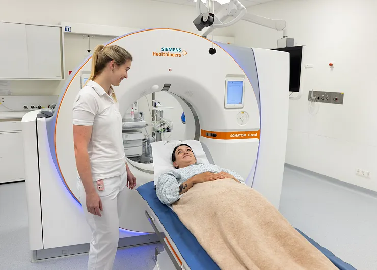 Eine medizinische Szenerie zeigt einen Patienten, der für ein CT-Scan auf einem Tisch liegt. Eine Krankenschwester steht lächelnd daneben. Der Raum ist modern.