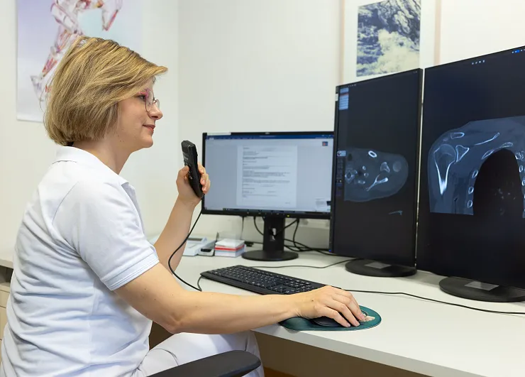 Eine medizinische Fachkraft in weißem Kittel betrachtet CT-Bilder auf zwei Monitoren und nutzt ein Handmikrofon zur Dokumentation oder Kommunikation.