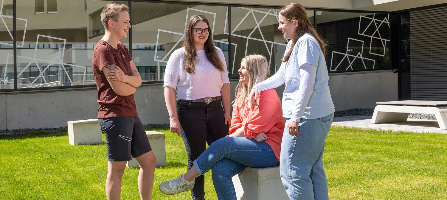 Eine Gruppe von vier jungen Erwachsenen unterhält sich fröhlich auf einer Grünfläche vor einem modernen Gebäude. Eine Person sitzt auf einem Betonblock.