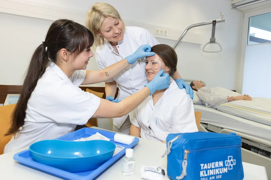 Zwei Ärztinnen in weißen Kitteln betreuen eine Patientin. Alle lächeln. Im Hintergrund liegt eine Patientin im Bett. Medizinische Ausrüstung liegt bereit.