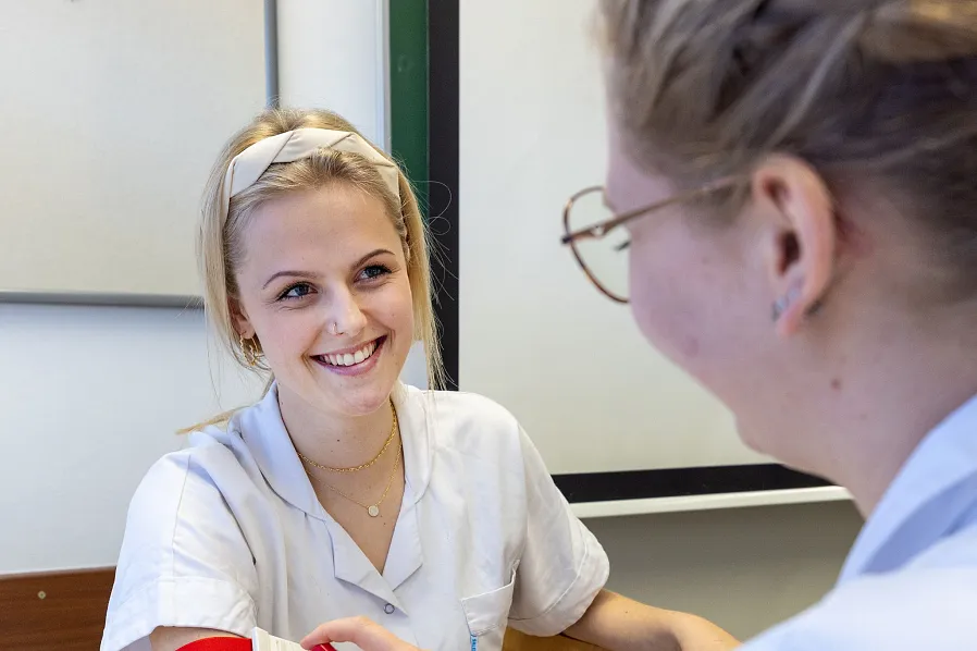 Zwei Personen in medizinischer Kleidung befinden sich in einem Behandlungsraum. Eine Person trägt ein Band um den Arm, während die andere ein Spray aufträgt.