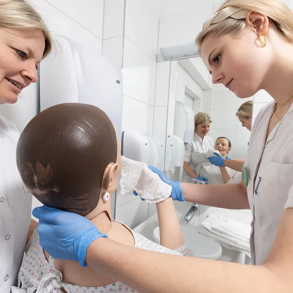 Zwei medizinische Fachkräfte in weißer Arbeitskleidung üben an einem realistischen Trainingsmodell in einem hellen, sterilen Raum, vielleicht in einer Klinik.