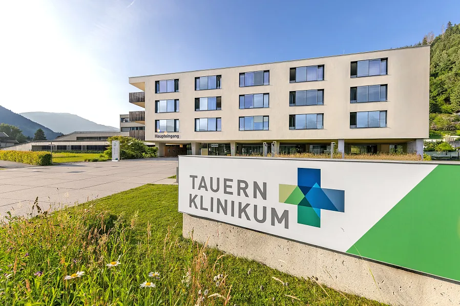 Modernes Gebäude in ländlicher Umgebung mit Schild "Tauernklinikum" im Vordergrund. Klare, blaue Himmel und grüne Wiese vervollständigen die Szene.