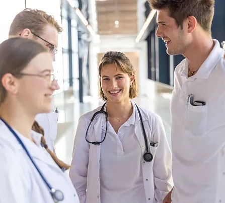 Eine Gruppe von fünf medizinischen Fachkräften in weißen Kitteln unterhält sich freundlich in einem modernen Klinikflur. Sie tragen Stethoskope.