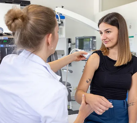 Eine Ärztin im weißen Kittel spricht mit einer Patientin, die auf einem Stuhl sitzt. Die Ärztin zeigt auf den Oberarm der Frau. Sie sind in einem Behandlungsraum.