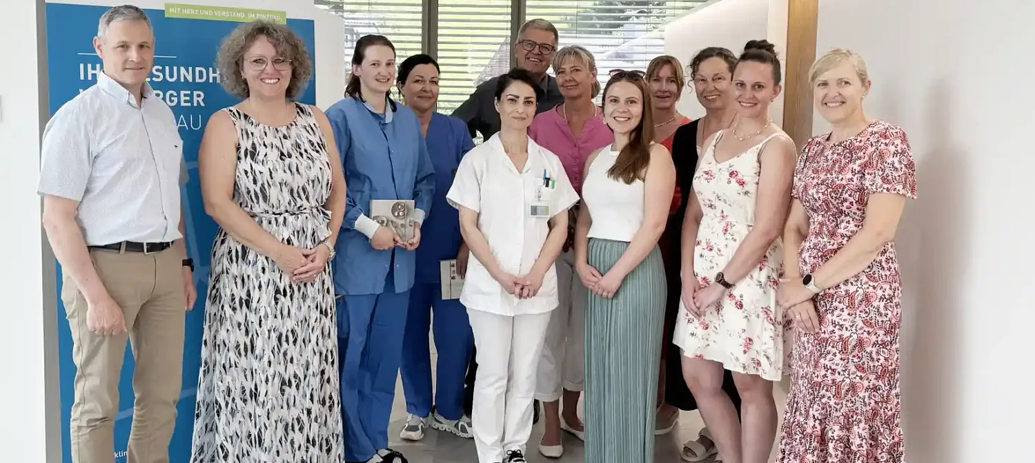 Eine Gruppe von medizinischem Personal und anderen Personen steht lächelnd in einem Raum. Sie tragen sowohl Freizeit- als auch Arbeitskleidung.