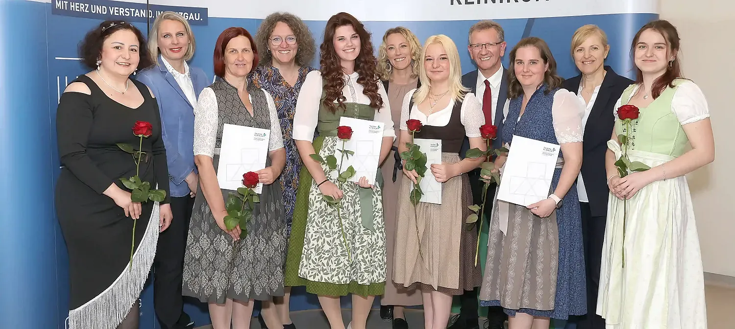 Eine Gruppe lächelnder Menschen in festlicher Kleidung posiert für ein Foto. Einige Personen halten rote Rosen und Urkunden. Sie stehen vor einem blauen Hintergrund.
