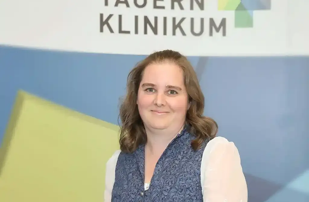 Eine Person mit braunen, schulterlangen Haaren steht vor einem Hintergrund mit dem Logo eines Klinikums. Sie trägt eine dunkelblaue Weste.