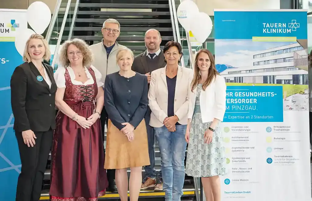 Eine Gruppe von sieben Personen steht vor einem Roll-Up mit der Aufschrift "Tauern Klinikum". Sie lächeln in die Kamera. Dahinter eine Treppe.
