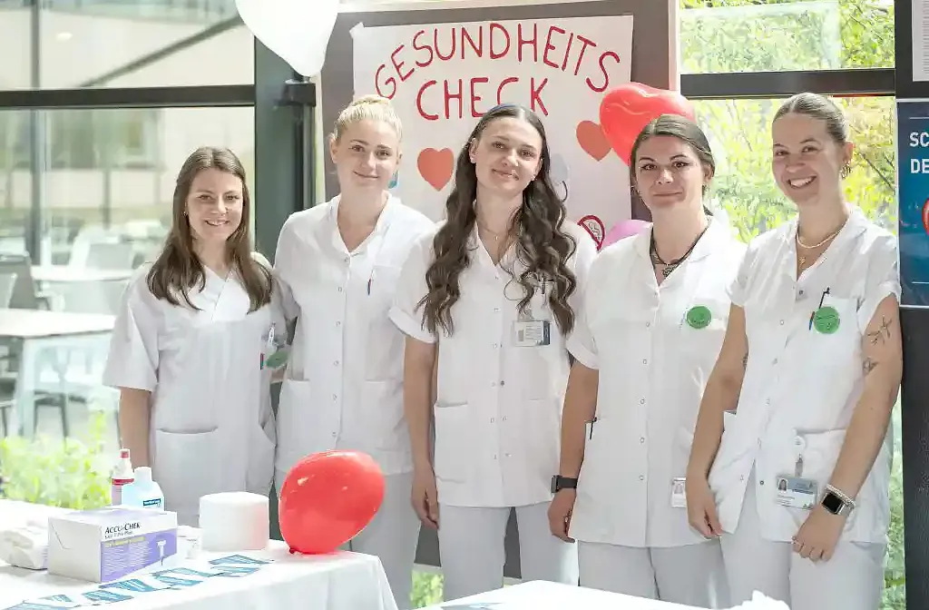 Fünf Personen in weißen Kitteln stehen lächelnd vor einem Gesundheitstisch mit roten Luftballons. Ein Schild im Hintergrund kündigt einen Gesundheitscheck an.