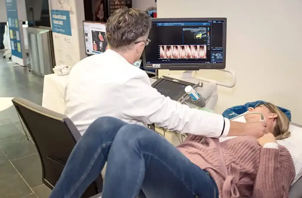 Ein Arzt untersucht eine Frau in einem Klinikraum mit Ultraschall auf einem Bildschirm. Sie liegt auf einer Liege und trägt eine Brille und einen rosa Pullover.