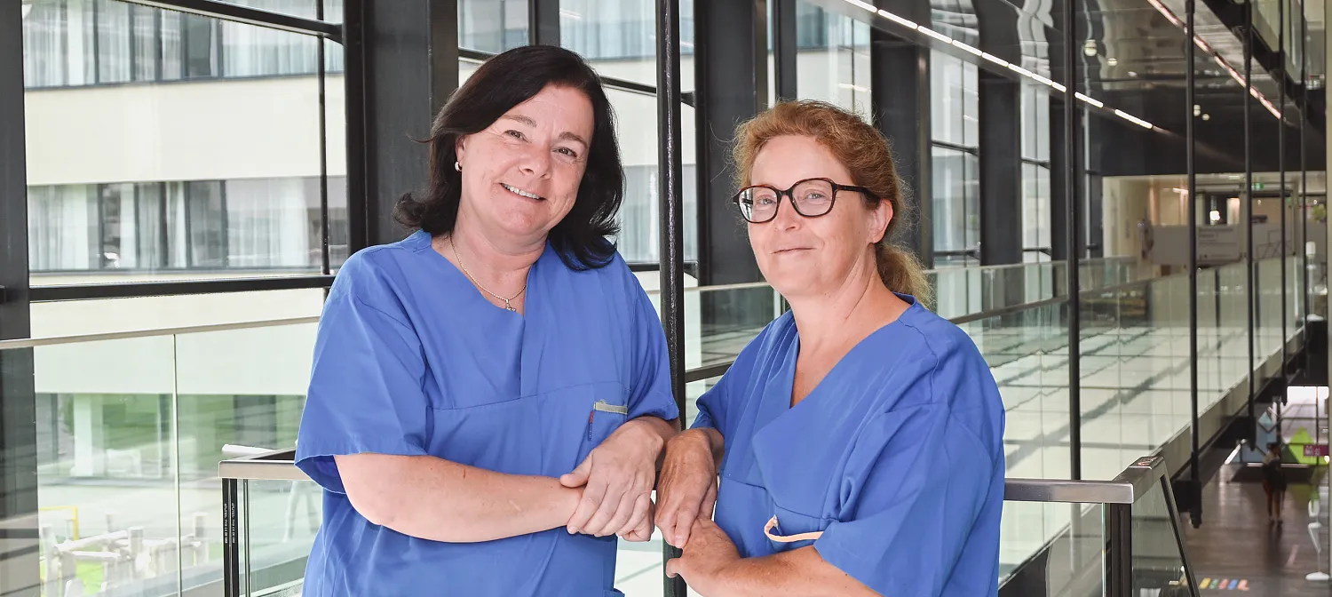 Zwei lächelnde Mitarbeiter:Innen in blauen medizinischen Uniformen stehen in einem modernen, verglasten Korridor eines Krankenhauses oder einer Klinik.