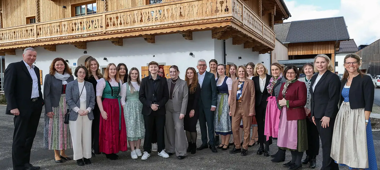 Eine Gruppe von Menschen steht in traditioneller und moderner Kleidung vor einem rustikalen Gebäude mit Holzverzierungen. Sie lächeln und posieren gemeinsam.