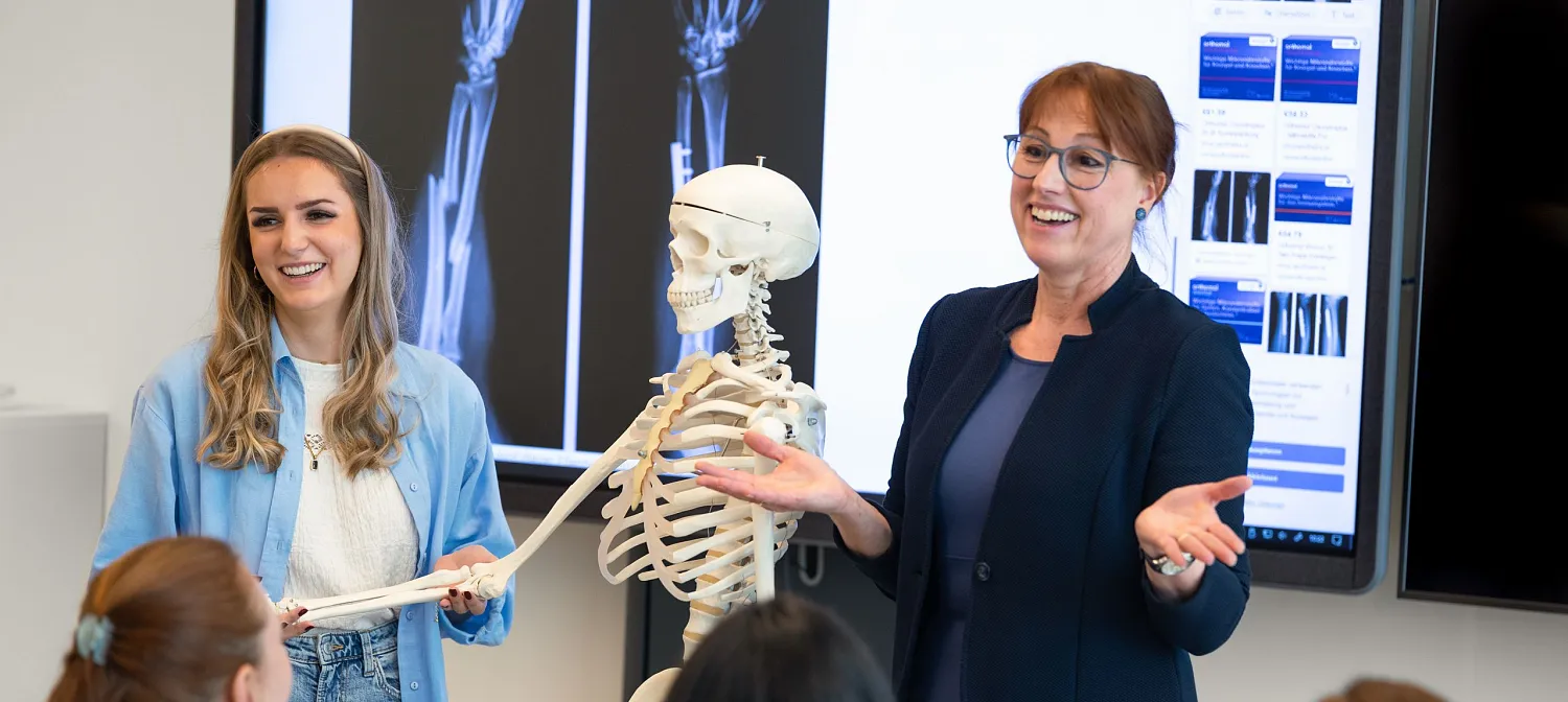 Zwei Frauen präsentieren vor Studierenden in einem Klassenraum. Ein Skelett und Röntgenbilder sind im Hintergrund zu sehen. Alle tragen lässige Kleidung.