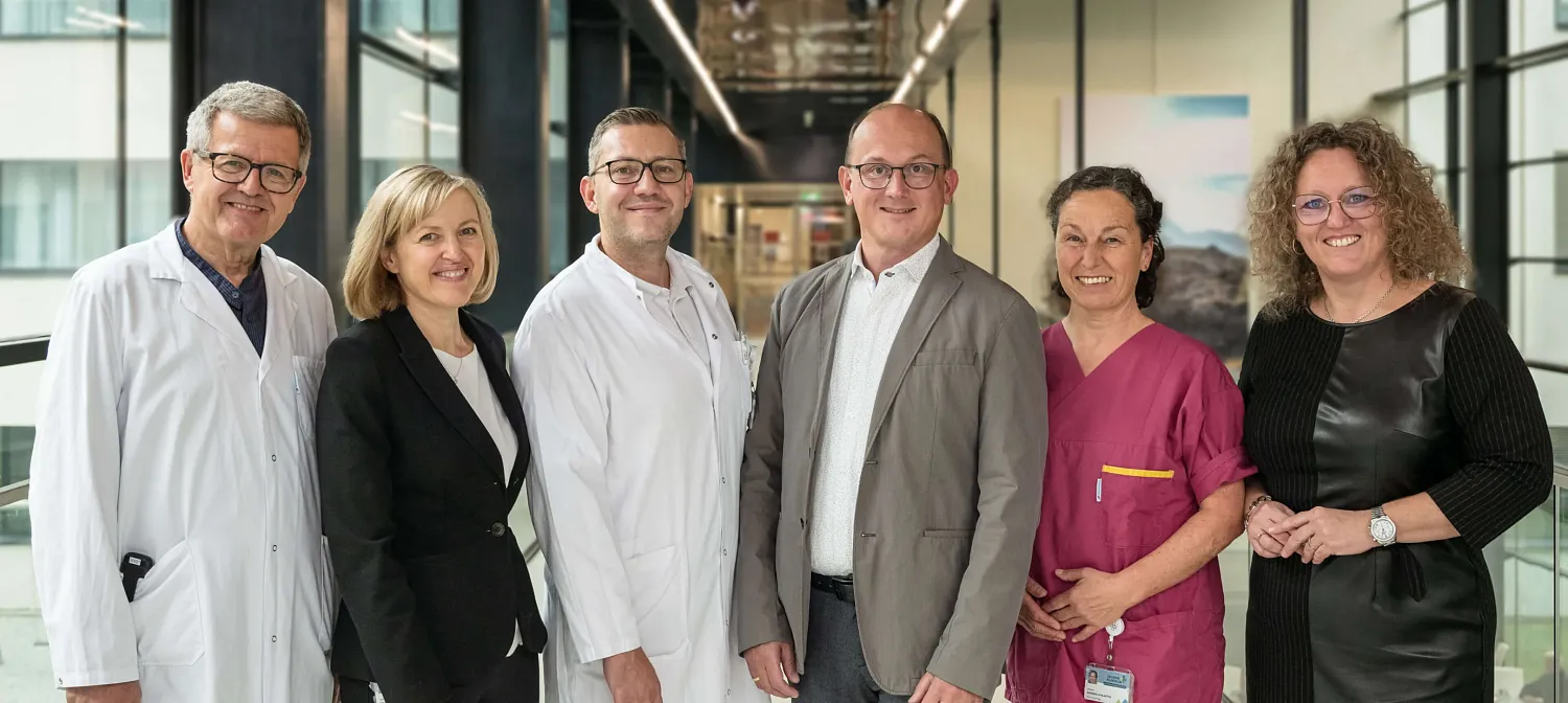 Sechs Personen, darunter Ärzte und medizinisches Personal, stehen lächelnd in einem modernen Krankenhausgang. Alle tragen professionelle Kleidung.