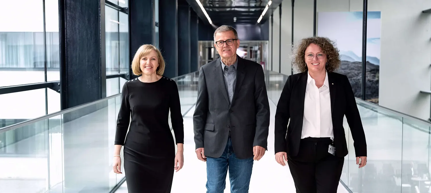 Drei Personen gehen lächelnd in einem modernen Bürogebäude einen hellen Flur entlang. Sie tragen Business-Kleidung und wirken professionell und freundlich.