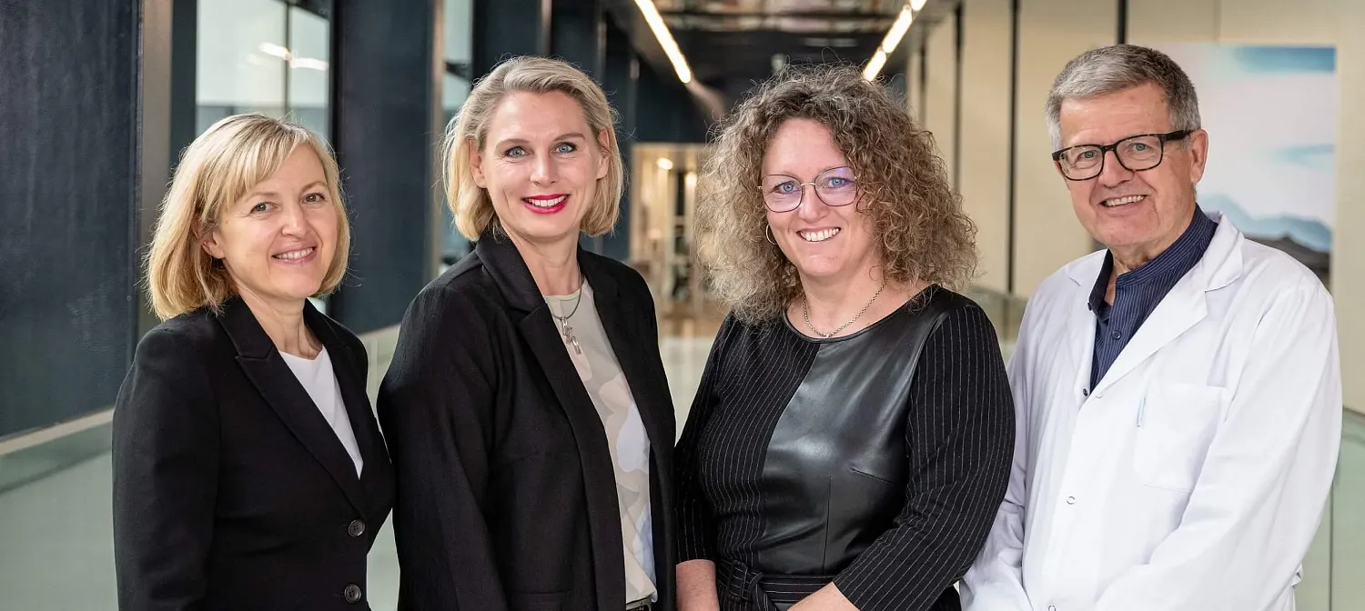 Gruppe von vier Personen, drei Frauen und ein Mann in einem Korridor. Sie tragen formelle Kleidung und lächeln in die Kamera. Der Hintergrund ist modern gestaltet.