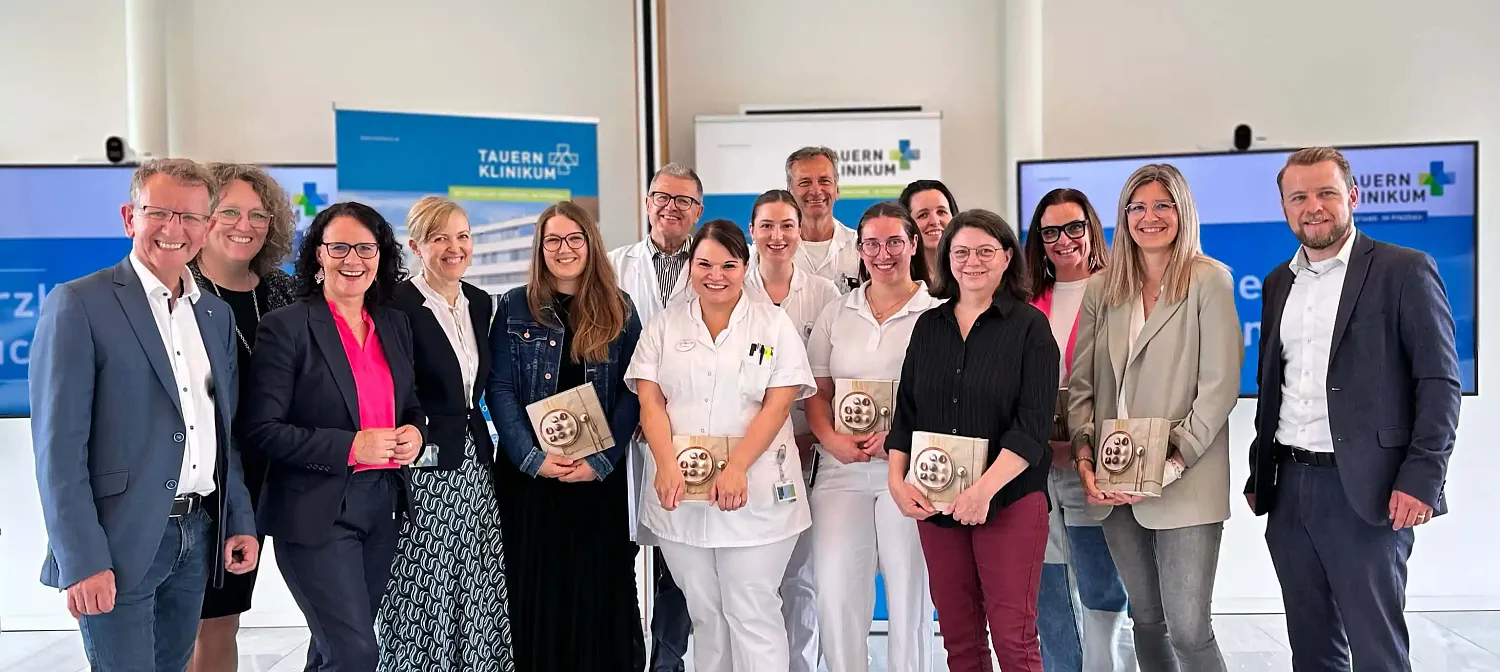 Eine Gruppe von Menschen steht lächelnd in einem Raum, einige tragen Krankenpflegekleidung. Im Hintergrund sind Krankenhausplakate sichtbar.