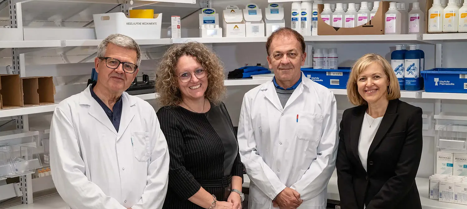 Gruppe von vier lächelnden Personen, zwei in weißen Laborkitteln, in einem Labor mit Regalen voller medizinischer Vorräte im Hintergrund.