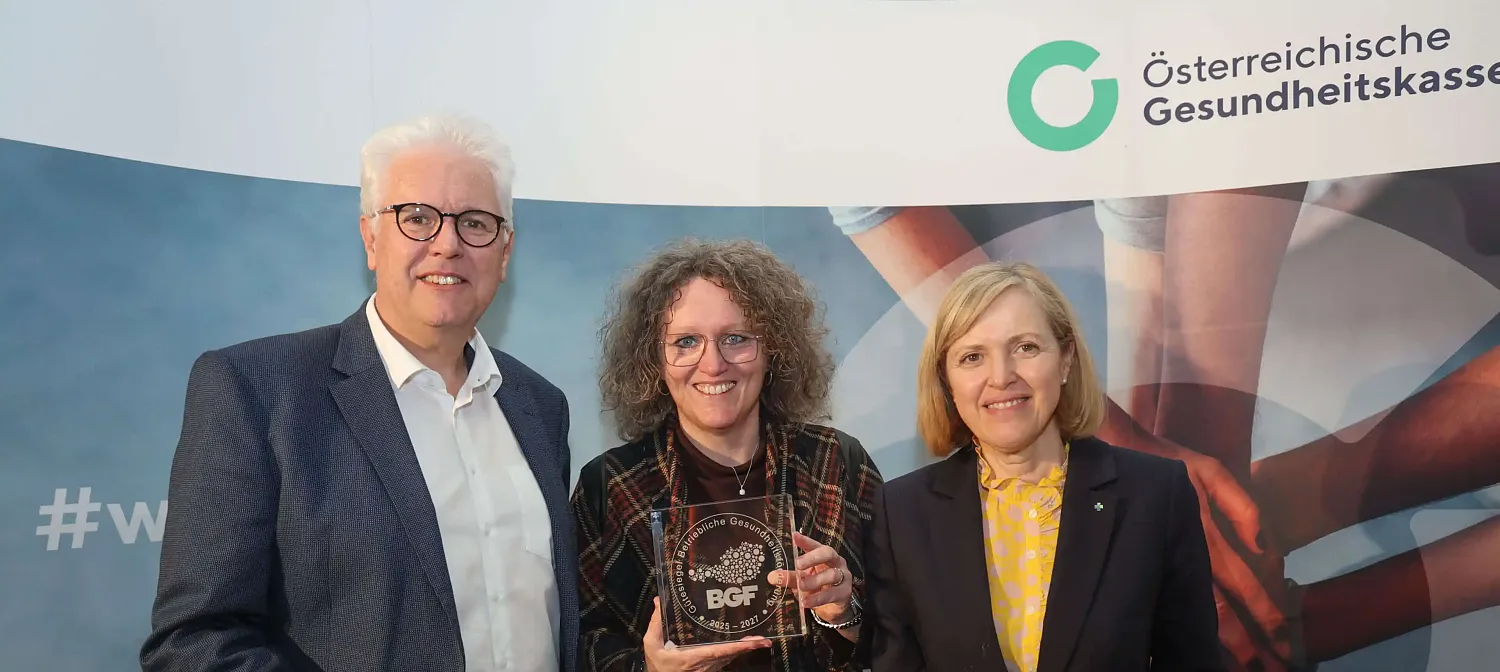 Drei Personen stehen vor einem Banner, eine hält eine Auszeichnung in den Händen. Sie lächeln alle in die Kamera. Der Hintergrund wirkt professionell.