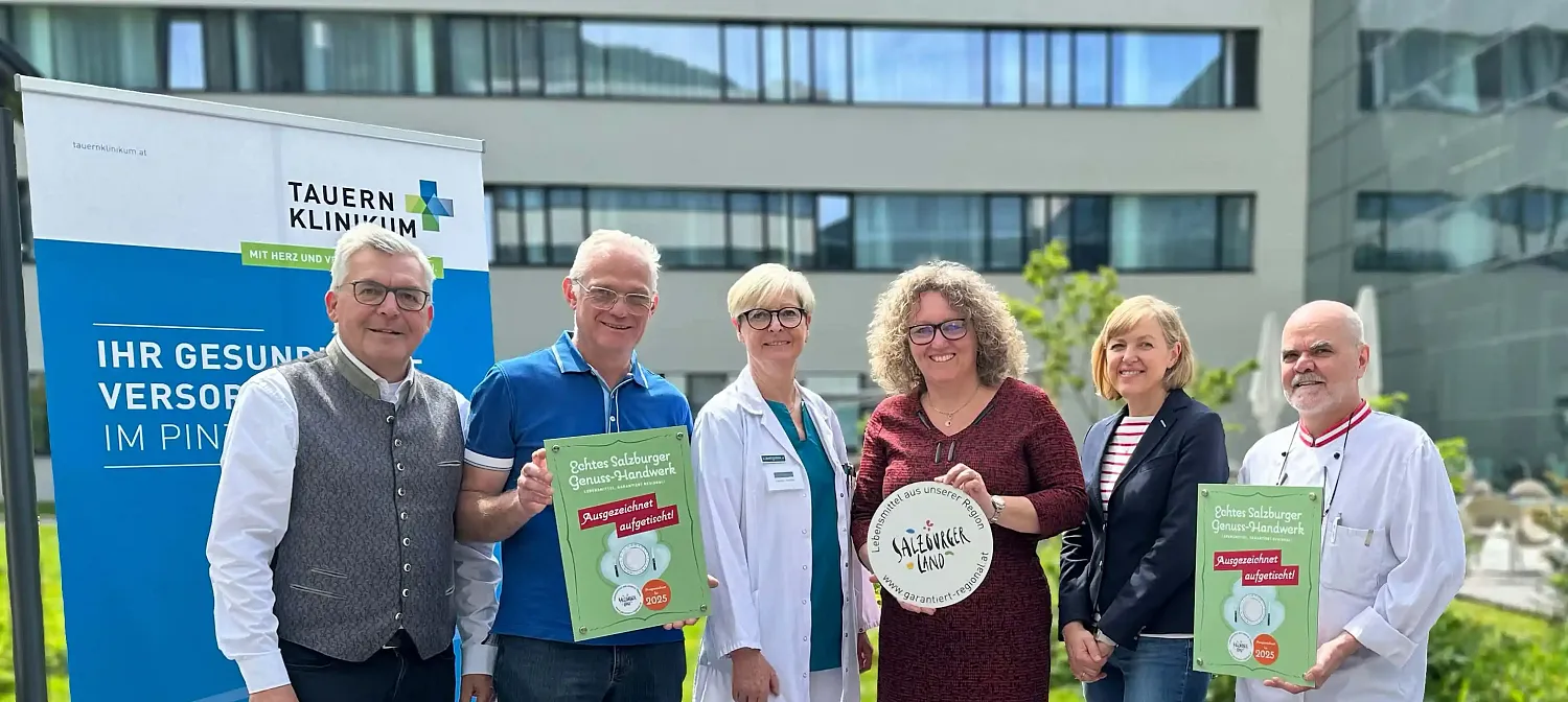 Eine Gruppe von sechs Personen, darunter Ärzte und Mitarbeitende eines Krankenhauses, posiert lächelnd mit einem Preis und Postern vor einem modernen Gebäude.