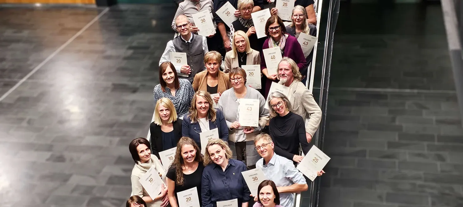 Eine Gruppe von Menschen steht in einer Treppe und hält Zertifikate in den Händen. Sie lächeln in die Kamera. Der Boden ist dunkelgrau gefliest.
