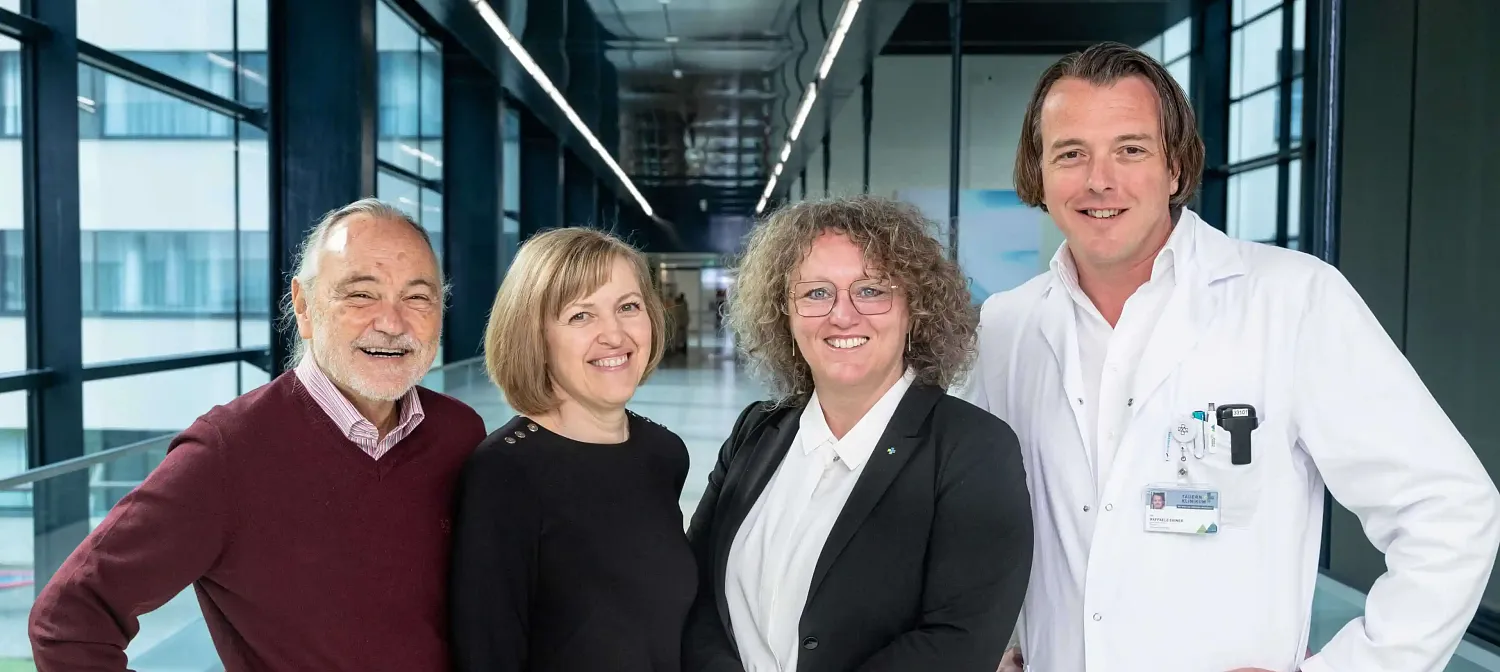 Eine Gruppe von vier Personen steht in einer modernen, hell beleuchteten Halle. Drei Frauen und ein Mann lächeln in die Kamera. Der Mann trägt einen Laborkittel.