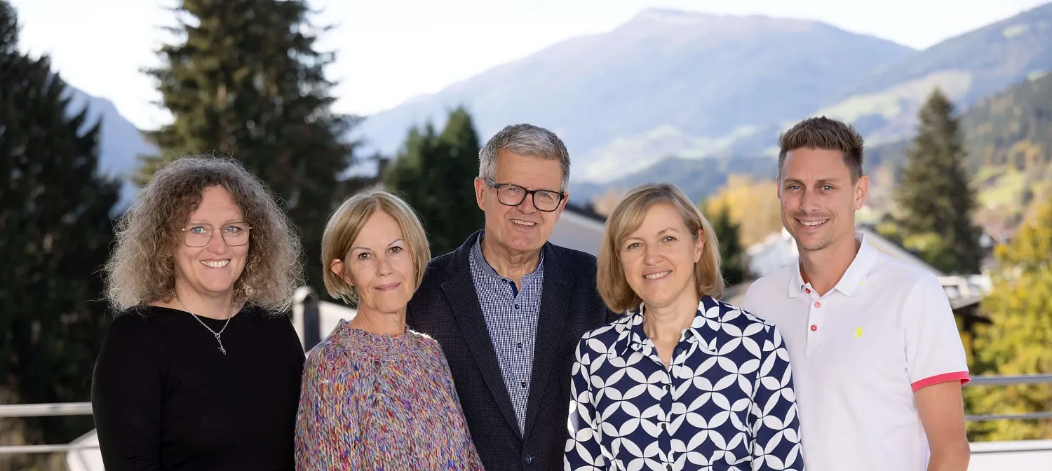 Fünf Personen stehen lächelnd im Freien vor einer bergigen Landschaft, hinter ihnen sind Bäume und Häuser sichtbar. Sie tragen unterschiedliche, farbenfrohe Kleidung.