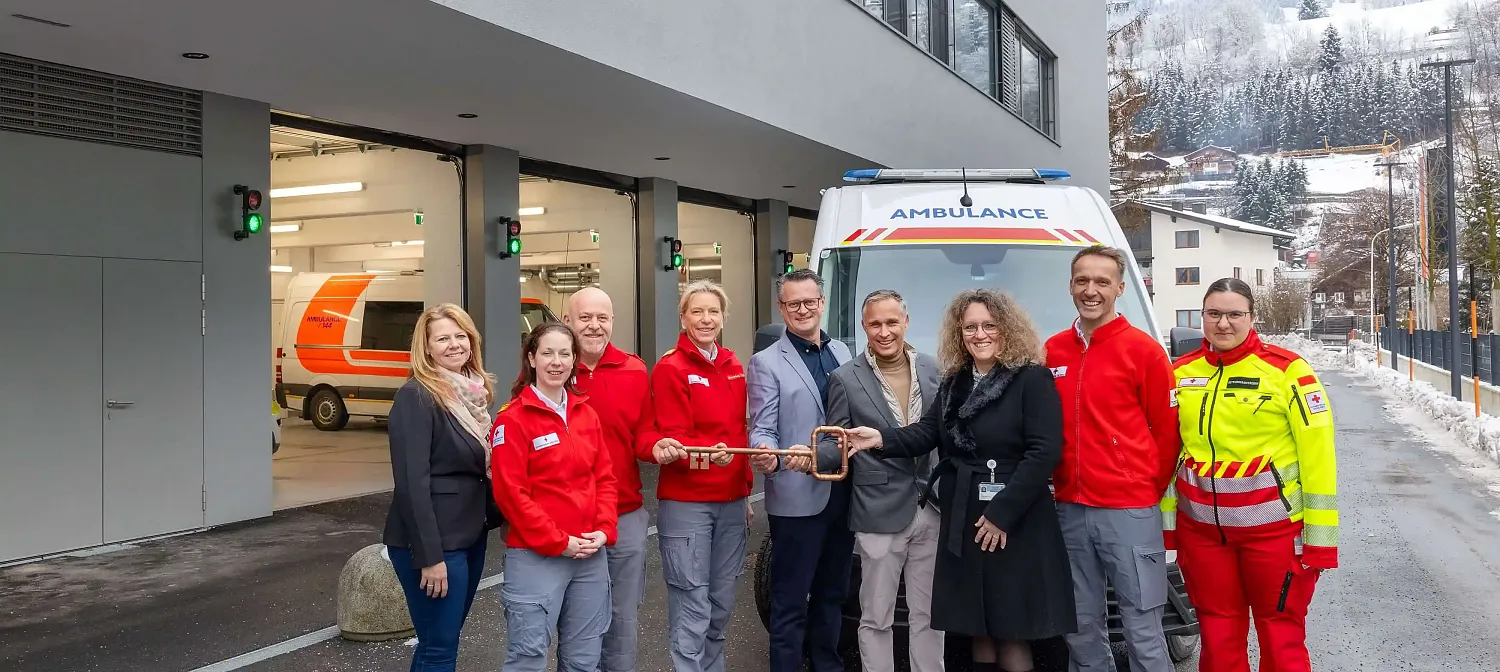 Eine Gruppe von Menschen steht vor einem Krankenwagen und einer Rettungsstation. Einige tragen rote Rettungsdienst-Uniformen, andere sind in Zivilkleidung.