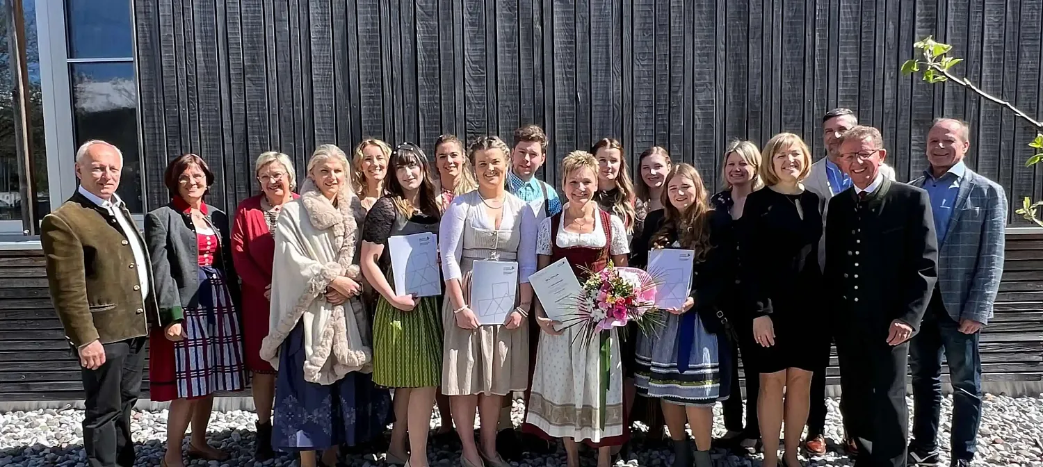 Eine Gruppe von Menschen in traditioneller bayerischer Tracht steht vor einer Holzwand. Einige Personen halten Urkunden und Blumensträuße in den Händen.