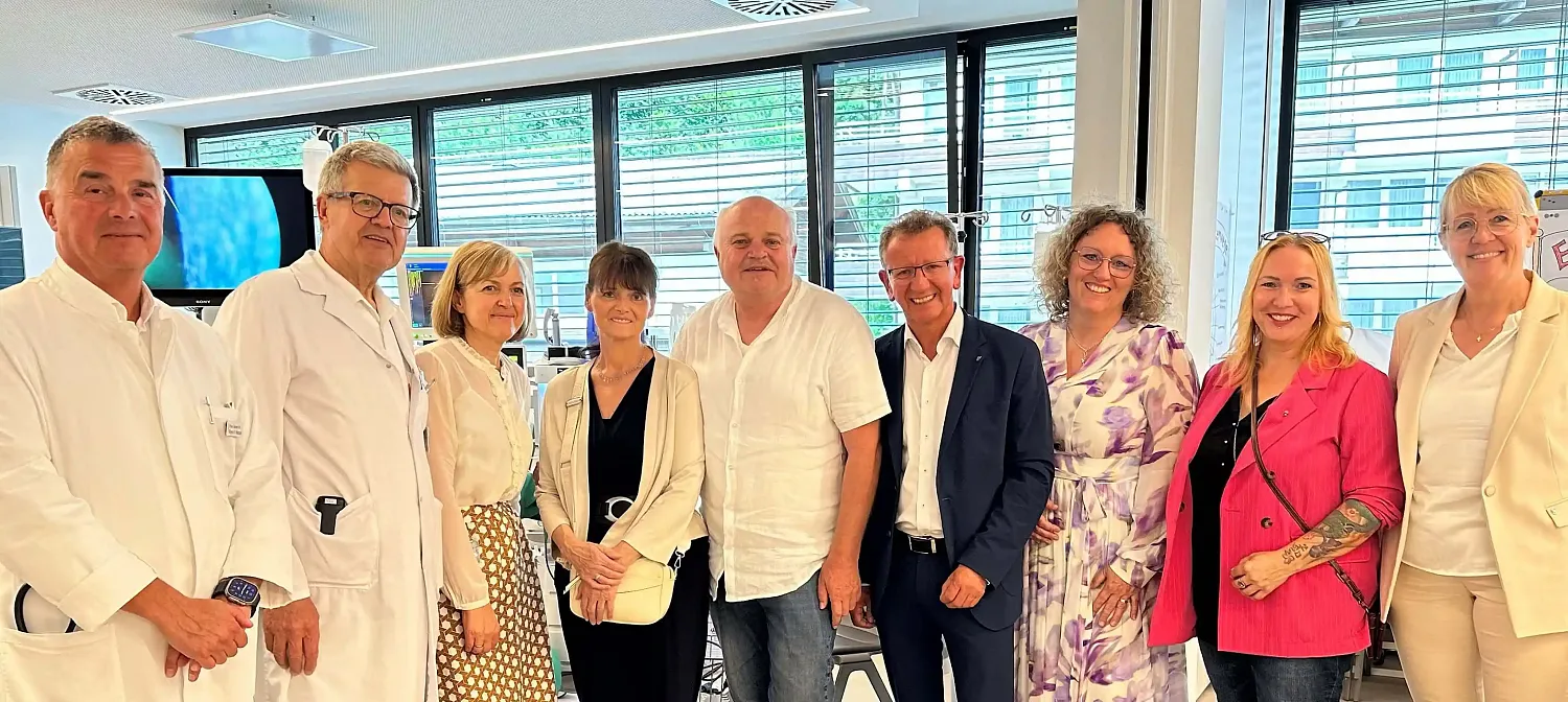 Eine Gruppe von sieben Personen steht nebeneinander in einem modernen Raum. Einige tragen formelle Kleidung, eine Person ist in einem Laborkittel.