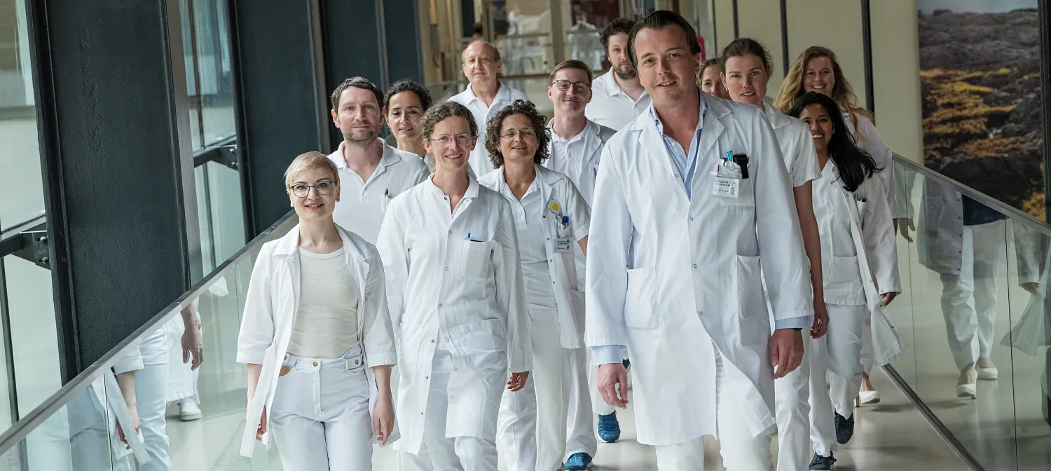 Eine Gruppe von Ärzten und Ärztinnen in weißen Kitteln geht lächelnd durch einen modernen Krankenhauskorridor mit Glaswänden und hellem Boden.