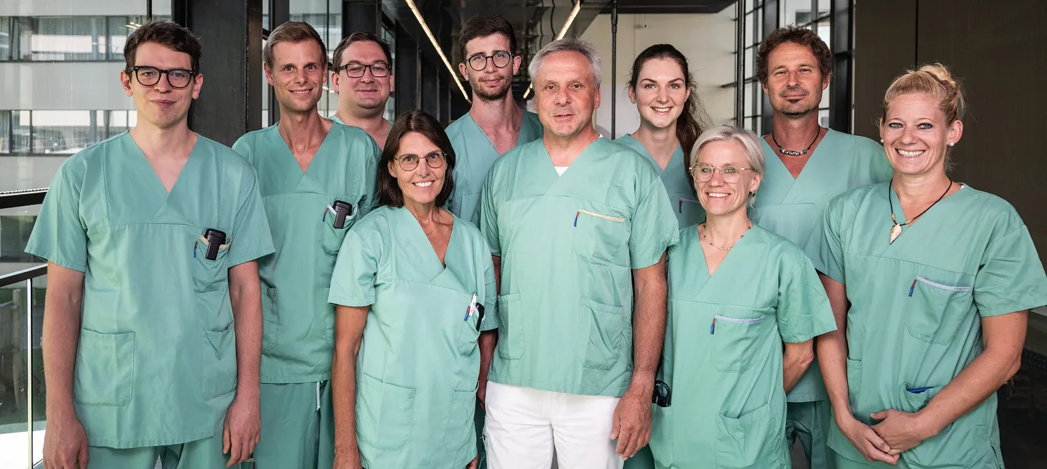 Eine Gruppe von medizinischem Personal in grüner OP-Kleidung steht zusammen und lächelt. Der Hintergrund zeigt einen modernen Krankenhausflur.