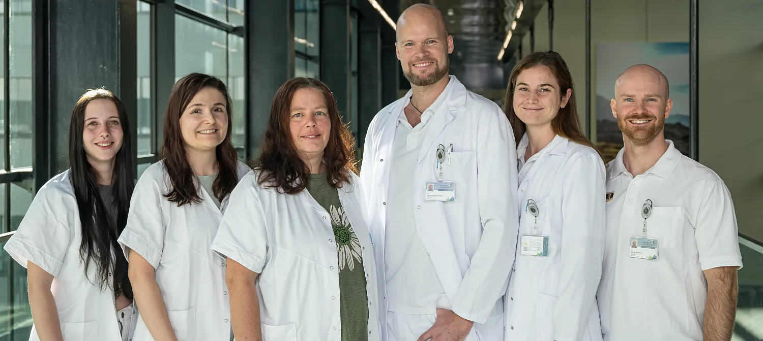 Eine Gruppe von sieben Personen in weißer Berufskleidung steht in einem modernen Gebäude mit Glaswänden und lächelt in die Kamera.