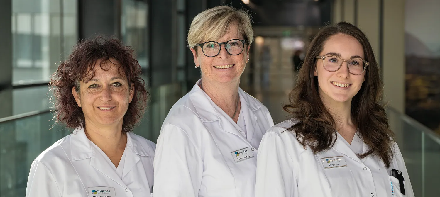 Drei Frauen in weißen Laborkitteln stehen lächelnd in einem modernen Gebäude. Sie tragen Namensschilder und Brillen und wirken professionell und freundlich.