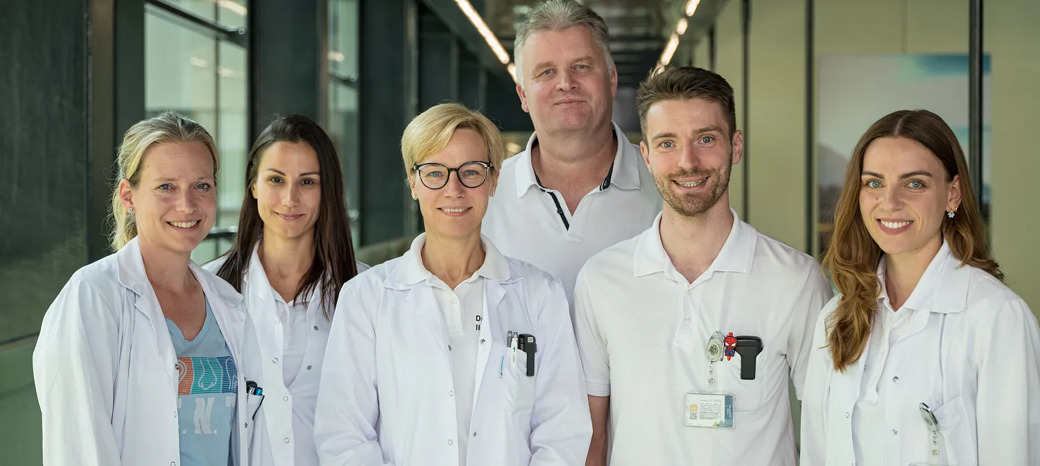 Eine Gruppe von sechs Personen in weißen medizinischen Kitteln steht in einem modernen Flur eines Krankenhauses. Sie lächeln und blicken in die Kamera.