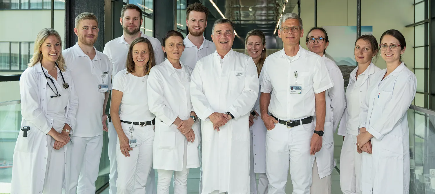A group of medical professionals in white coats stand in a hospital corridor, smiling and posing for a group photo. The setting is bright and modern.