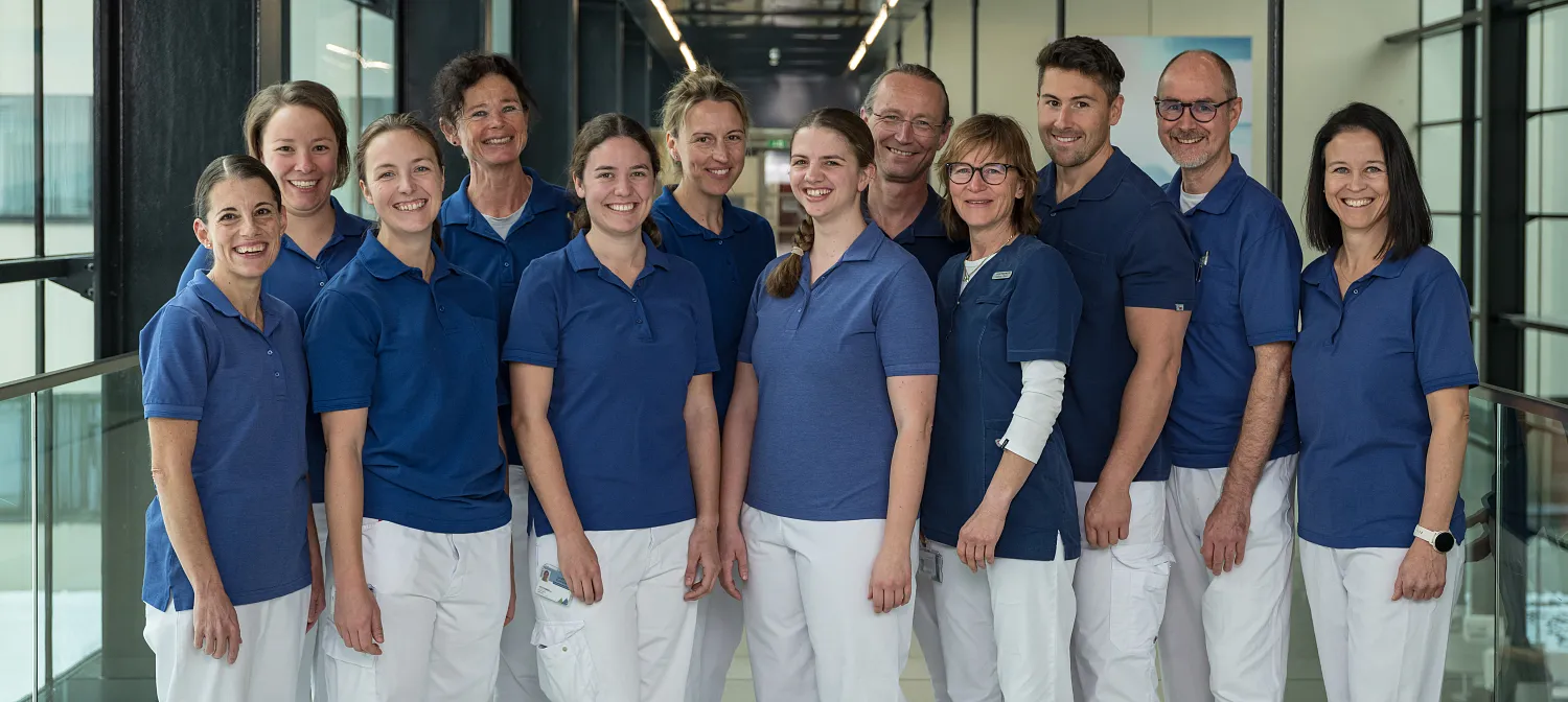 Eine Gruppe von zwölf medizinischen Fachleuten in blauen und weißen Uniformen steht lächelnd auf einem modernen, gut beleuchteten Flur im Krankenhaus.