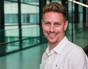 Ein lächelnder Mann in einem weißen Hemd steht vor einem modernen Glasgebäude mit großen Fenstern. Der Hintergrund ist unscharf, was den Fokus auf ihn legt.