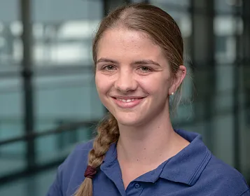 Eine lächelnde Frau in einem blauen Poloshirt mit geflochtenem Haar steht vor einer Glaswand in einem modernen Gebäude. Sie wirkt zufrieden und freundlich.