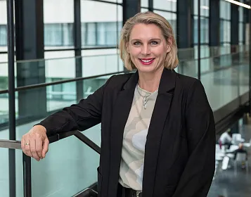 Eine lächelnde Frau mit blonden Haaren steht in einem modernen, verglasten Gebäude an einer Metallgeländer. Sie trägt einen schwarzen Blazer und ein gemustertes Shirt.