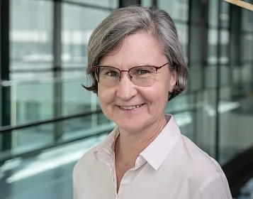 Porträt einer lächelnden Person mittleren Alters mit grauem Haar und Brille, in einem modernen, verglasten Gebäude.