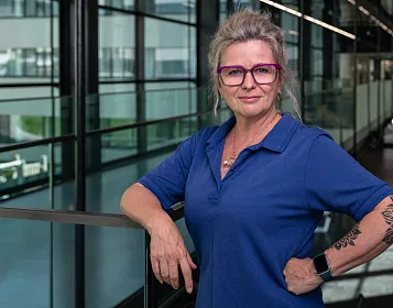 Eine Person mit Brille und blauer Kleidung steht in einem modernen gläsernen Korridor. Außenbereich und Architektur spiegeln sich in den Fenstern wider.