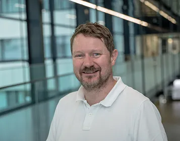 Ein lächelnder Mann in einem weißen Polohemd steht in einem modernen Gebäude mit großen Fenstern. Der Hintergrund zeigt eine verschwommene Innenarchitektur.